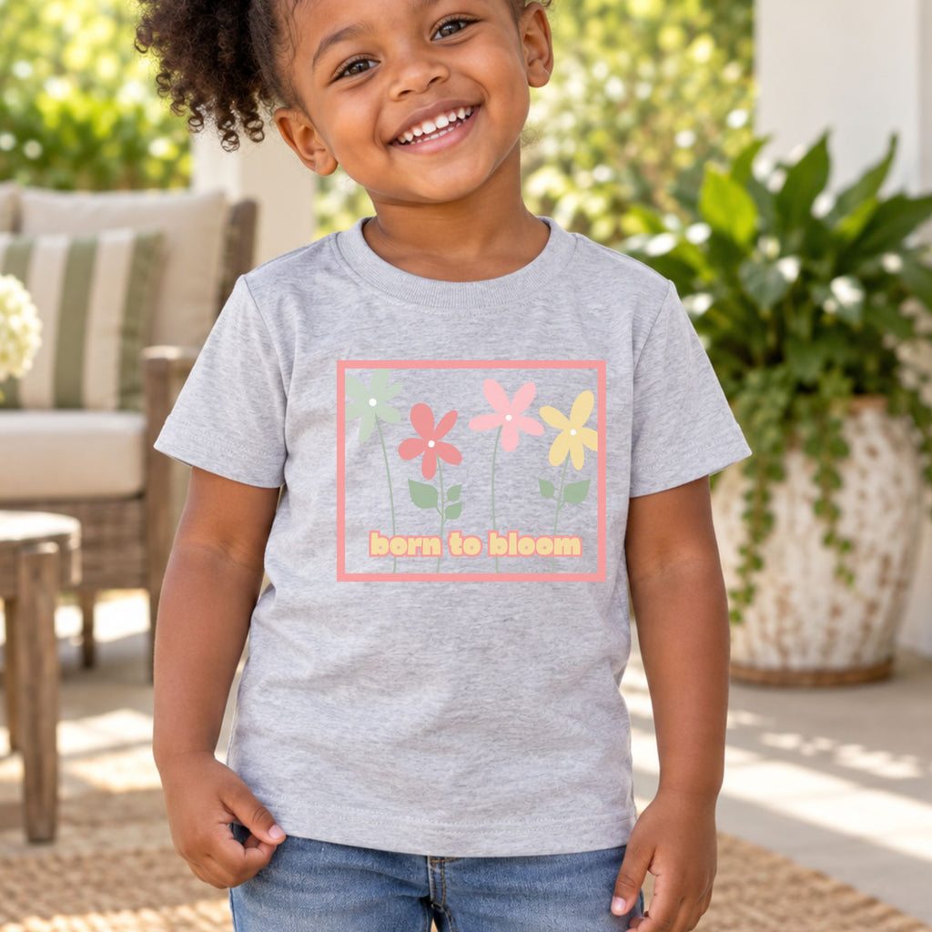 Toddler wearing a heather gray Born to Bloom short sleeve tee on a porch, front view showing the colorful retro flower design.