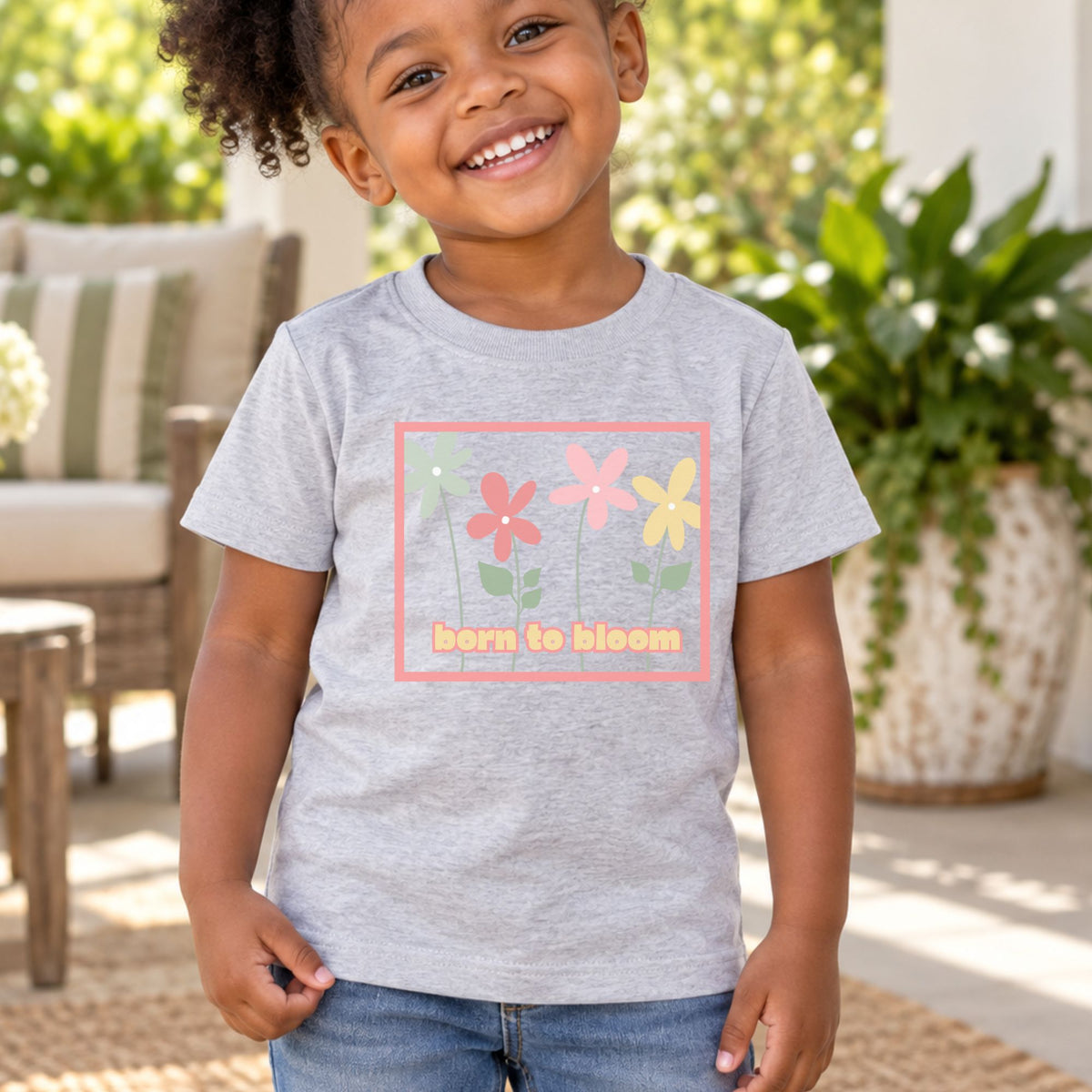 Toddler wearing a heather gray Born to Bloom short sleeve tee on a porch, front view showing the colorful retro flower design.