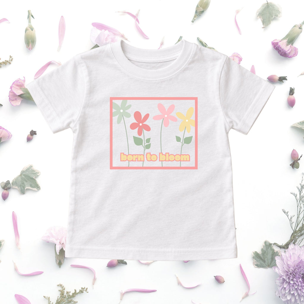 White Born to Bloom toddler tee displayed in a floral flat lay, showing the colorful retro flower graphic.