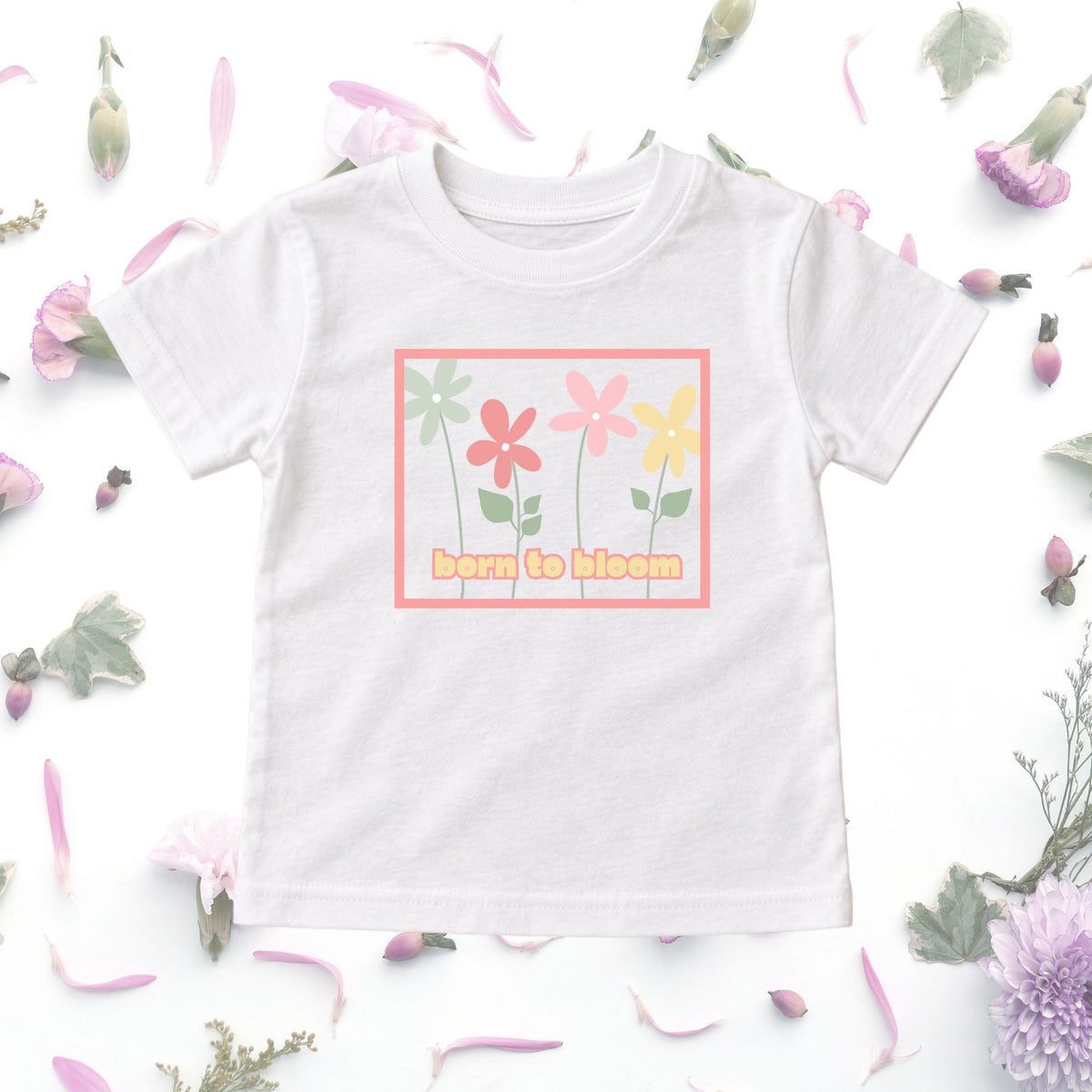 White Born to Bloom toddler tee displayed in a floral flat lay, showing the colorful retro flower graphic.