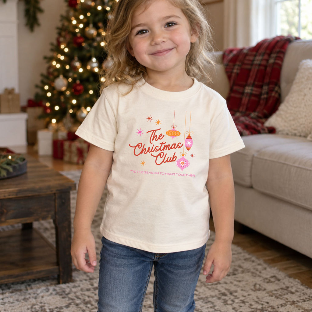 Toddler wearing a natural Christmas Club short sleeve tee in a festive living room, front view showing the retro ornament design and Tis the Season to Hang Together message.