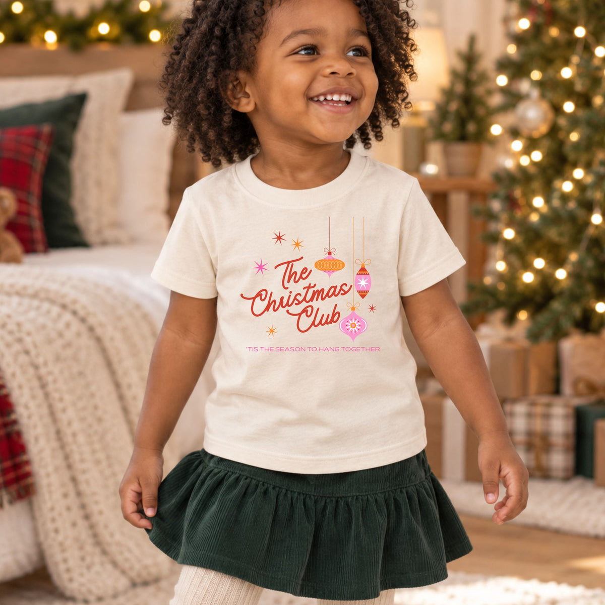 Toddler wearing a natural Christmas Club short sleeve tee in a holiday setting, front view showing the retro ornament design and Tis the Season to Hang Together message.
