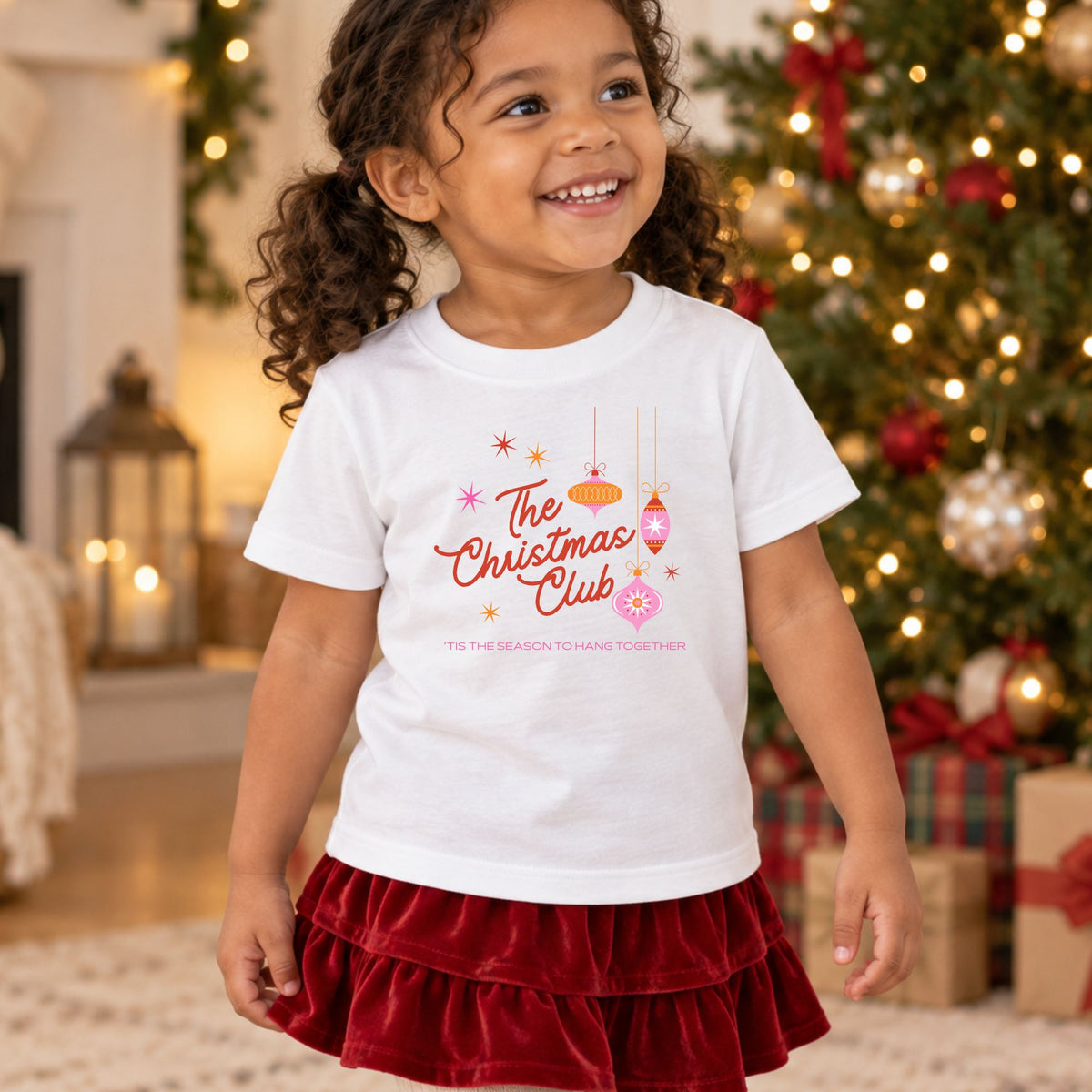 Toddler wearing a white Christmas Club short sleeve tee by a Christmas tree, front view showing the retro ornament design and Tis the Season to Hang Together message.
