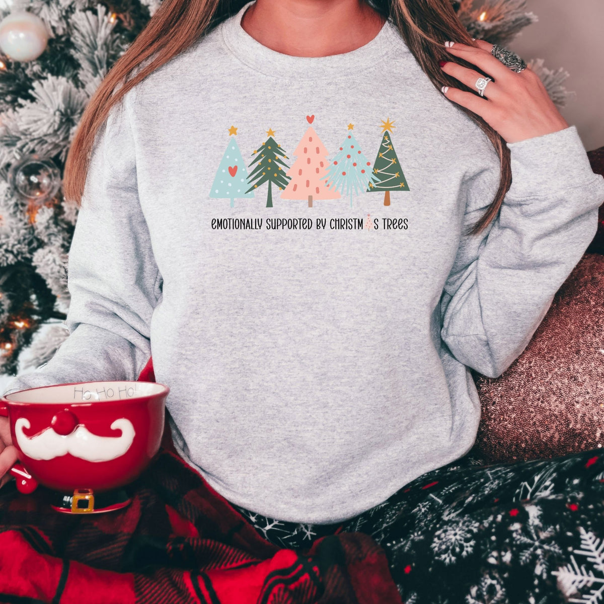 Woman wearing an ash gray Emotionally Supported by Christmas Trees adult crewneck sweatshirt beside a Christmas tree.