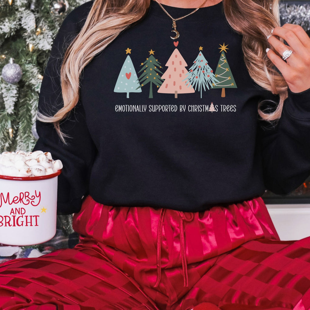 Woman wearing a black Emotionally Supported by Christmas Trees adult crewneck sweatshirt with red holiday pajamas.