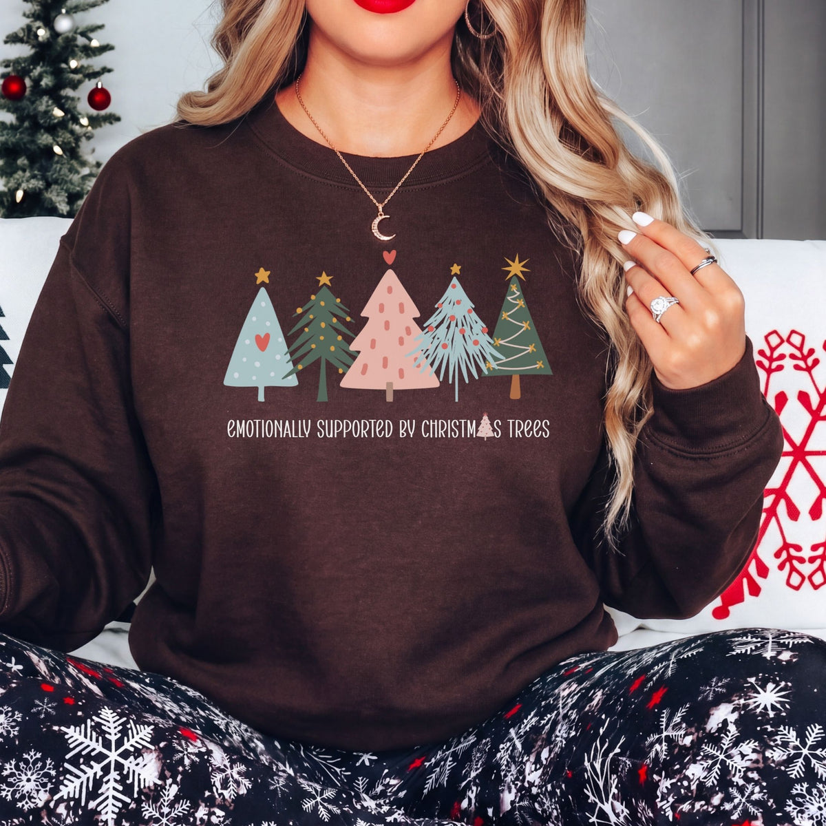 Woman wearing a dark brown Emotionally Supported by Christmas Trees adult crewneck sweatshirt with holiday pajamas.