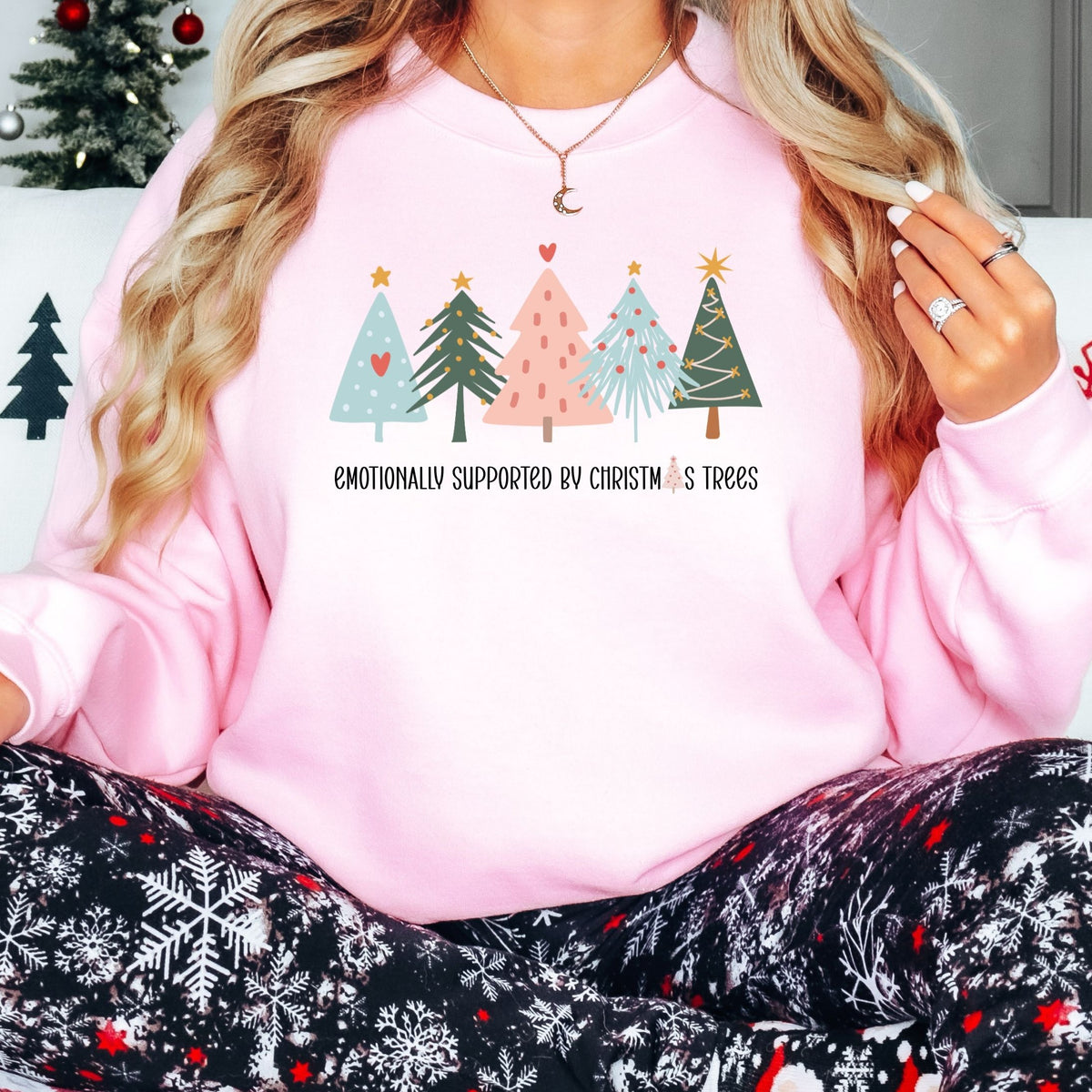 Woman wearing a light pink Emotionally Supported by Christmas Trees adult crewneck sweatshirt with holiday pajamas.