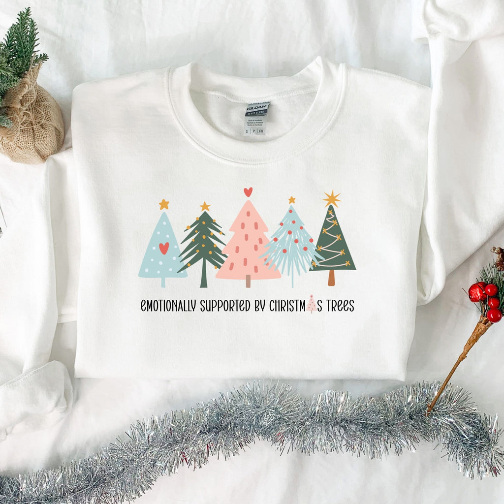 Folded white Emotionally Supported by Christmas Trees adult crewneck sweatshirt in a Christmas flat lay.