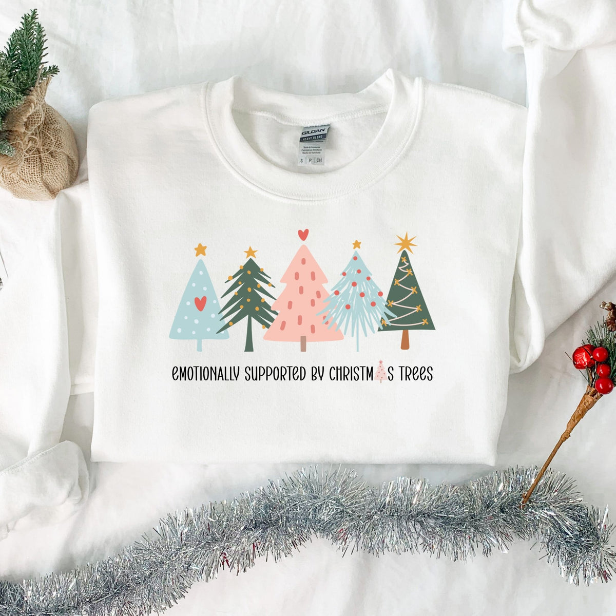 Folded white Emotionally Supported by Christmas Trees adult crewneck sweatshirt in a Christmas flat lay.