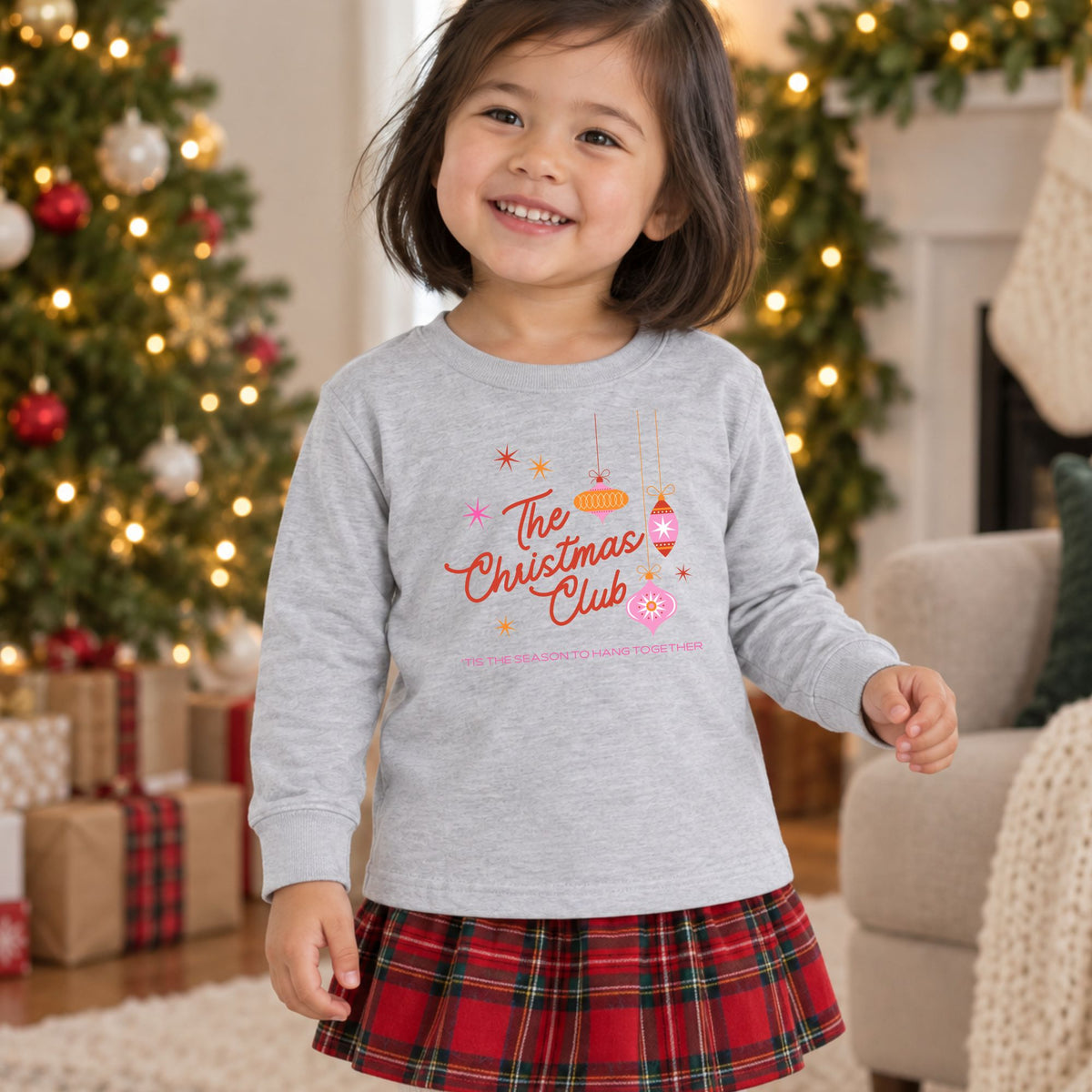 Girl wearing a heather gray The Christmas Club toddler long sleeve tee in a Christmas setting, showing the colorful retro ornament front graphic.