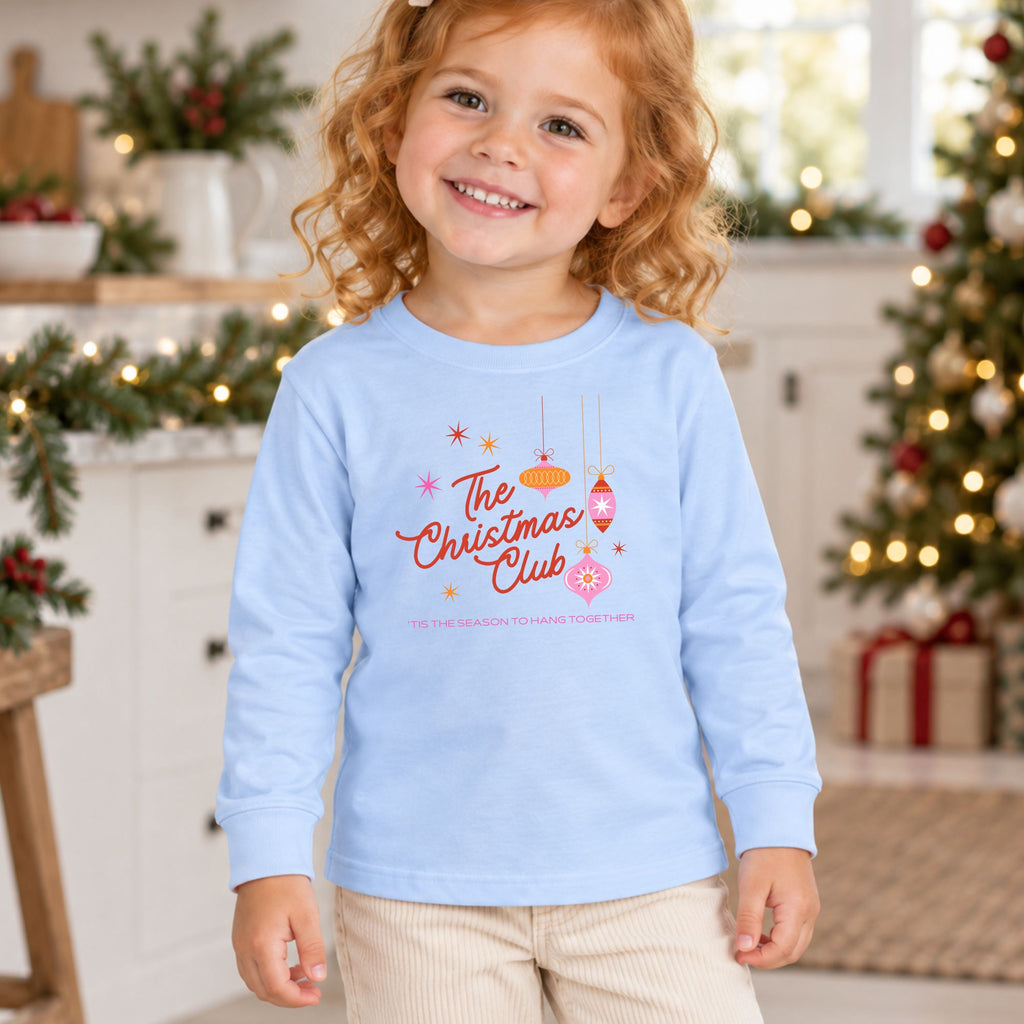 Girl wearing a light blue The Christmas Club toddler long sleeve tee in a Christmas setting, showing the colorful retro ornament front graphic.