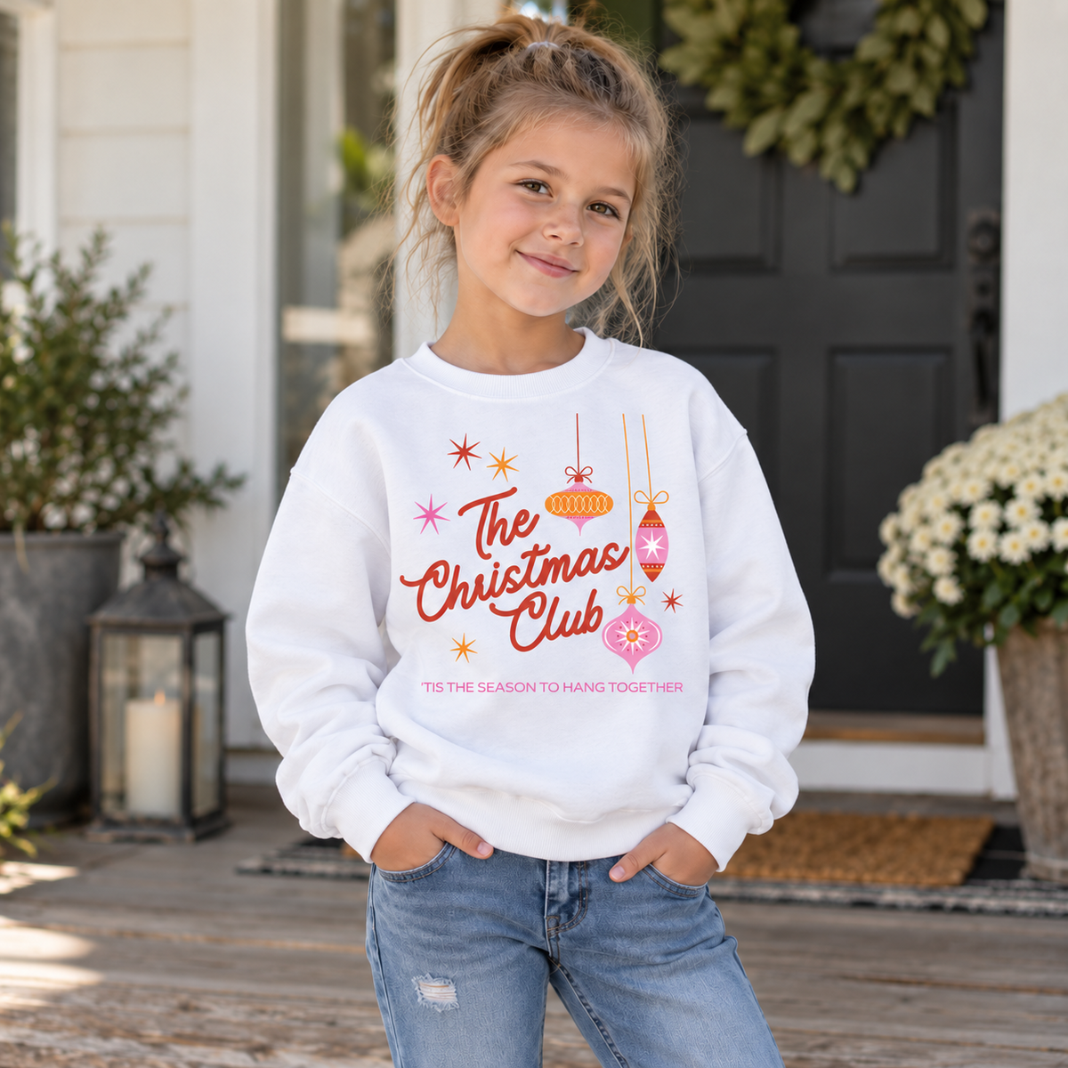 Girl wearing a white The Christmas Club ’Tis the Season to Hang Together youth crewneck sweatshirt on a porch.